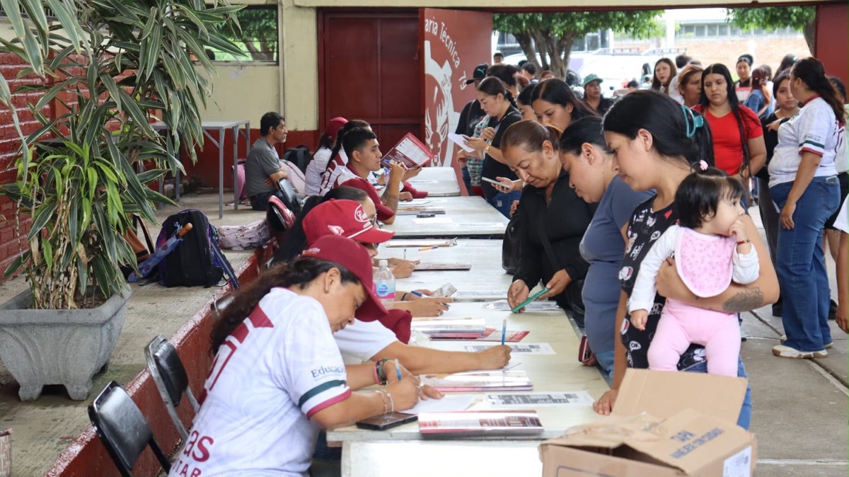 Varias personas hacen fila y completan formularios en mesas atendidas por jóvenes con gorras y camisetas, mientras una mujer sostiene a un bebé.