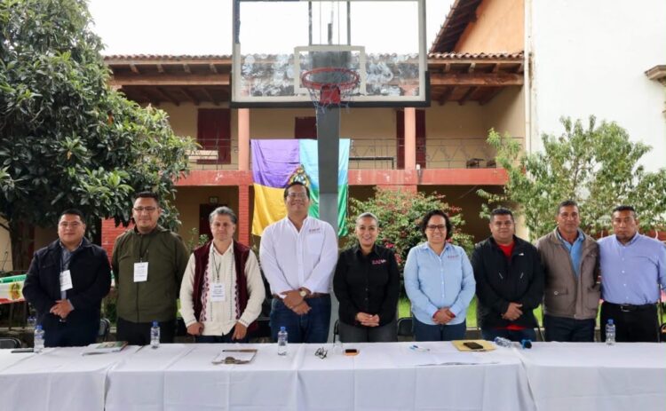 Un grupo de nueve personas está de pie detrás de una mesa larga al aire libre, con un aro de baloncesto y un edificio de fondo.