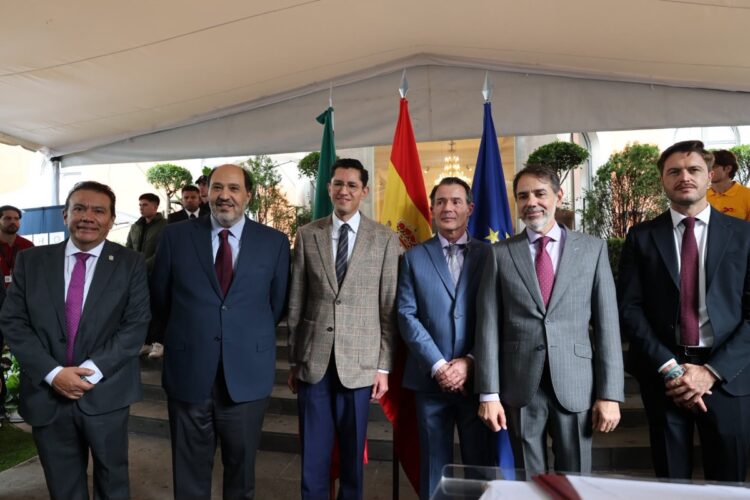 Seis hombres con trajes formales posan juntos bajo una carpa, con banderas de México, España y la Unión Europea detrás de ellos.