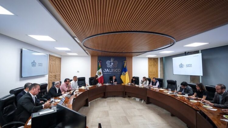 Varias personas están sentadas alrededor de una mesa de conferencias en una sala de reuniones con pantallas y banderas.