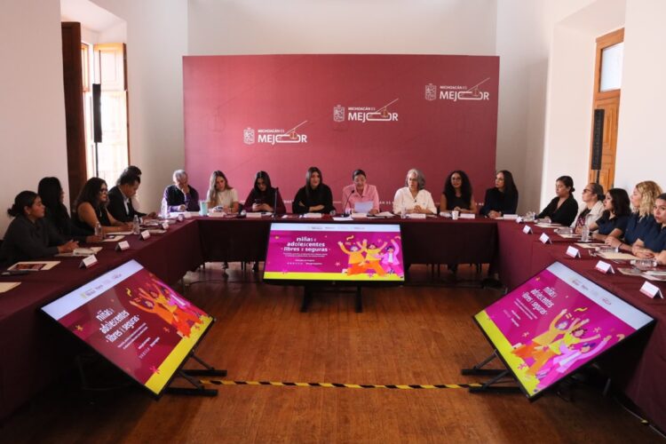 Un grupo de personas está sentado alrededor de una mesa en forma de U en una sala de reuniones con pantallas y un fondo rojo.