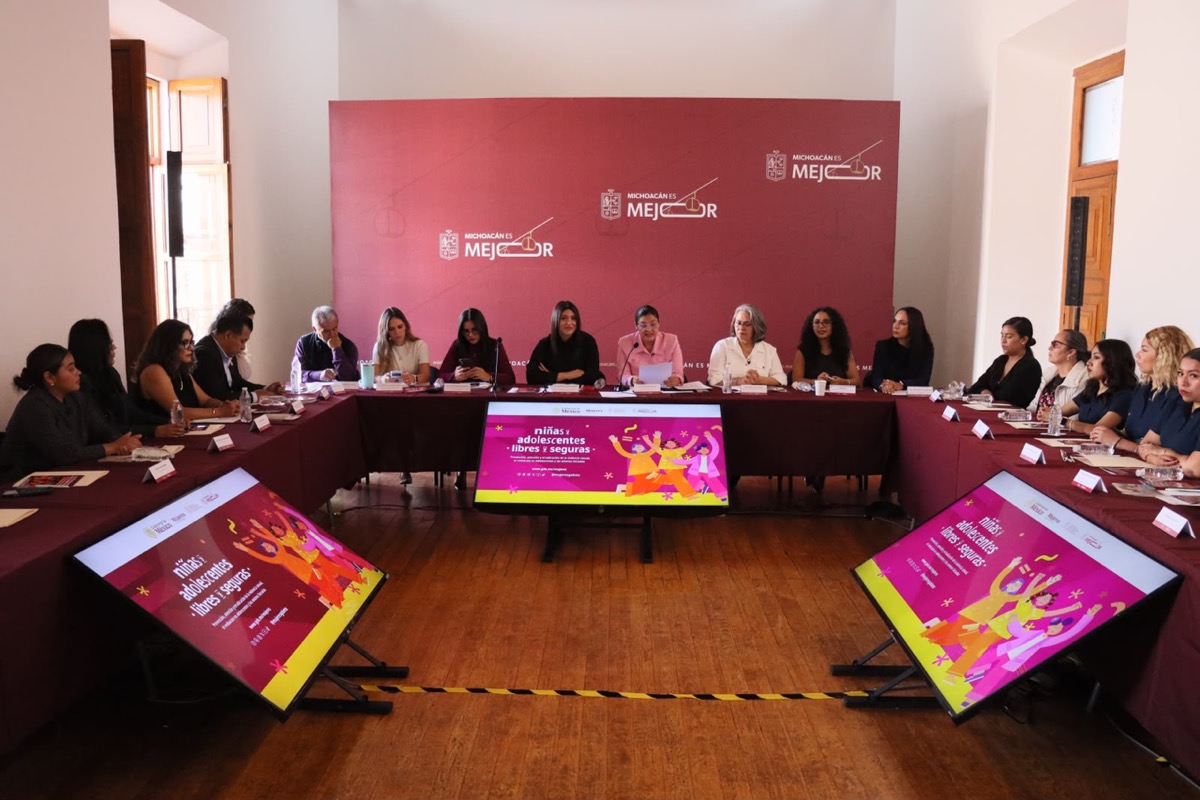 Un grupo de personas está sentado alrededor de una mesa en forma de U en una sala de reuniones con pantallas y un fondo rojo.