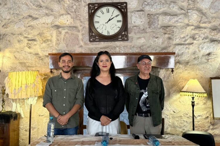 Tres personas están de pie detrás de una mesa con botellas de agua, frente a una pared de piedra con un reloj y dos lámparas.