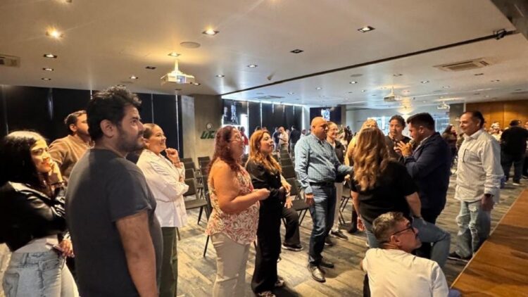 Un grupo de personas está reunido en una sala, de pie y conversando entre sillas vacías.