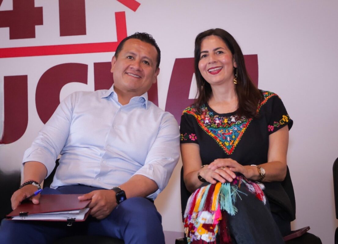 Un hombre y una mujer sonríen mientras están sentados juntos frente a un fondo con letras rojas.
