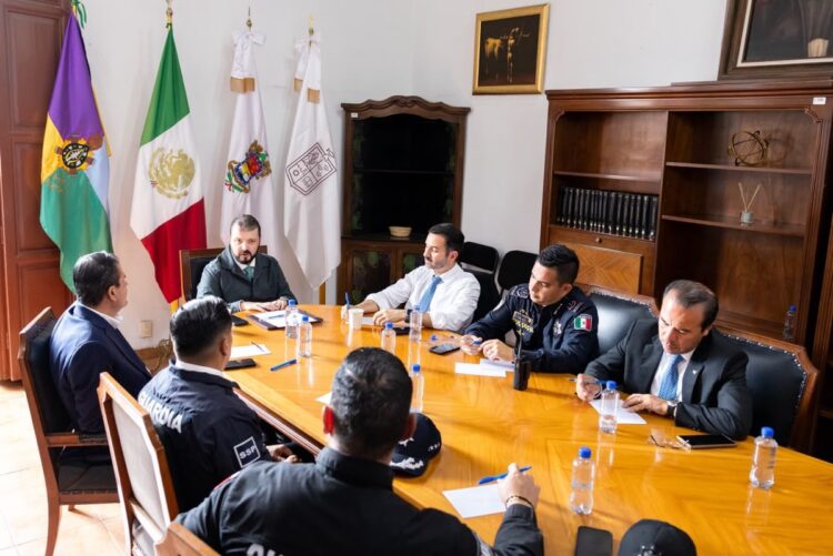 Varias personas, incluyendo oficiales de policía, están reunidas alrededor de una mesa de madera en una sala con banderas y estanterías.