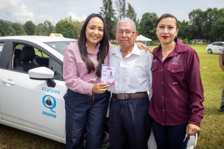 Tres personas sonríen frente a un taxi blanco en un campo, mientras una de ellas sostiene una credencial.