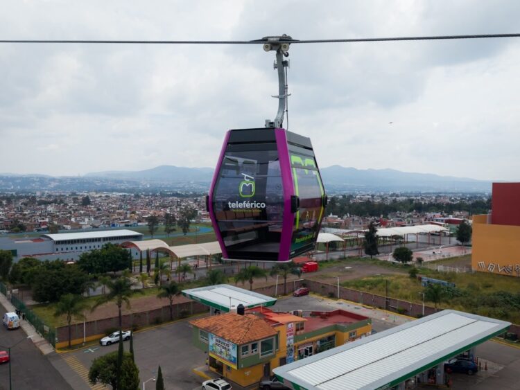 Un teleférico morado y negro se desplaza sobre una ciudad con montañas al fondo.