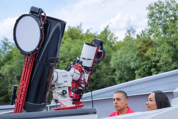 Dos personas observan un telescopio grande al aire libre rodeado de árboles.