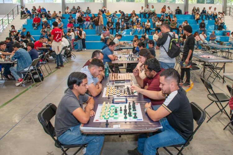 Varias personas juegan ajedrez en un torneo dentro de un gimnasio con público sentado en gradas azules.