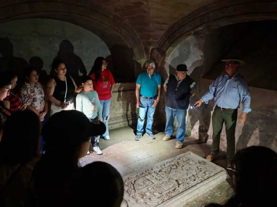 Grupo de personas observando una lápida o relieve tallado en piedra dentro de una bóveda, con iluminación tenue y sombras de征.