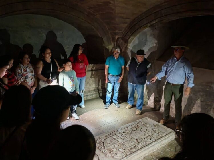 Un grupo de personas observa una tumba de piedra iluminada por una linterna en una cripta oscura.