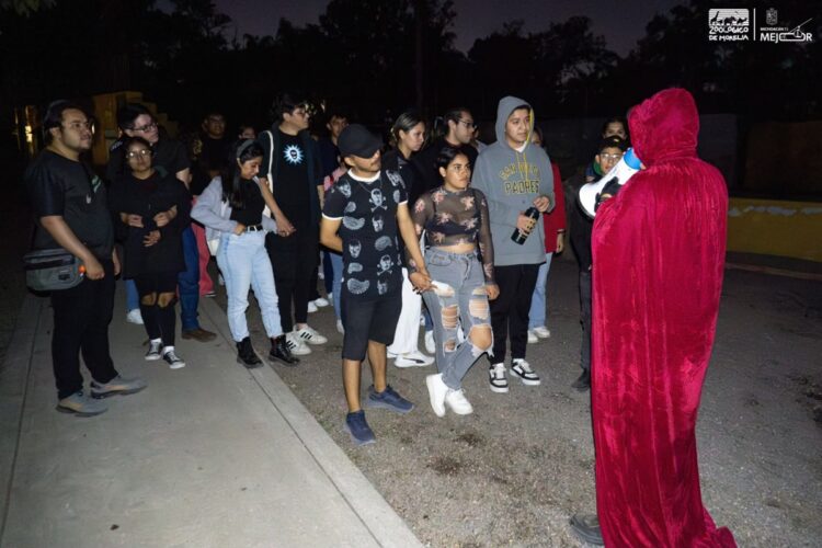 Un grupo de personas escucha a alguien con capa roja y megáfono durante la noche en un espacio al aire libre.