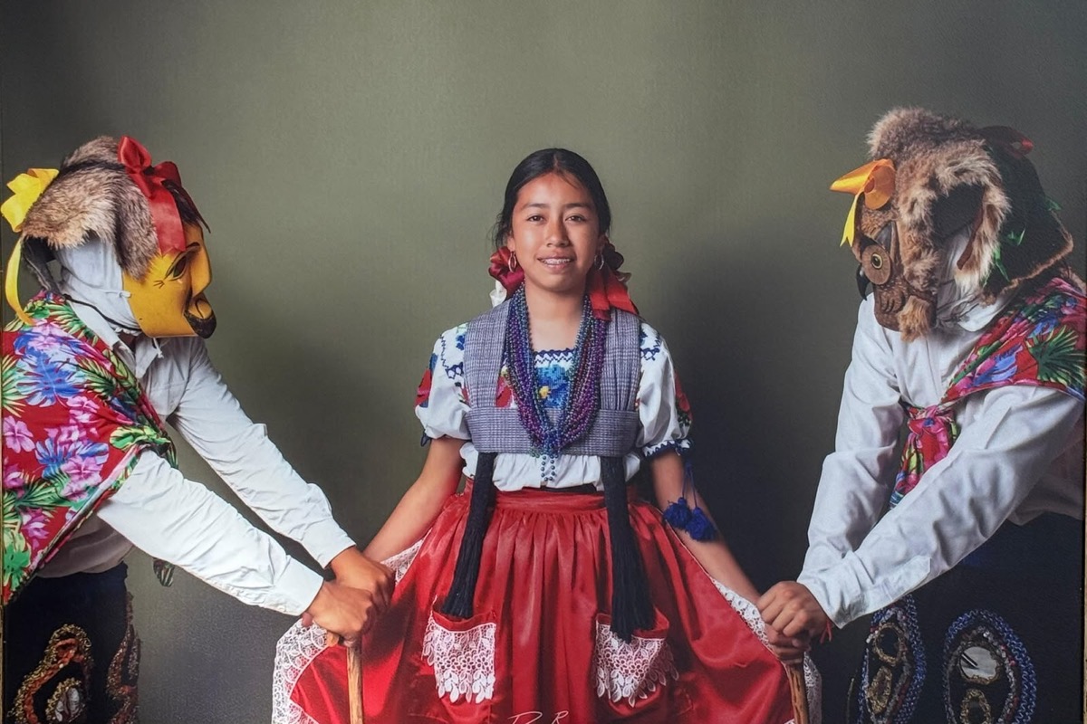 Tres personas con trajes tradicionales, dos con máscaras de animales y una niña en el centro con vestido rojo, posan frente a un fondo liso.