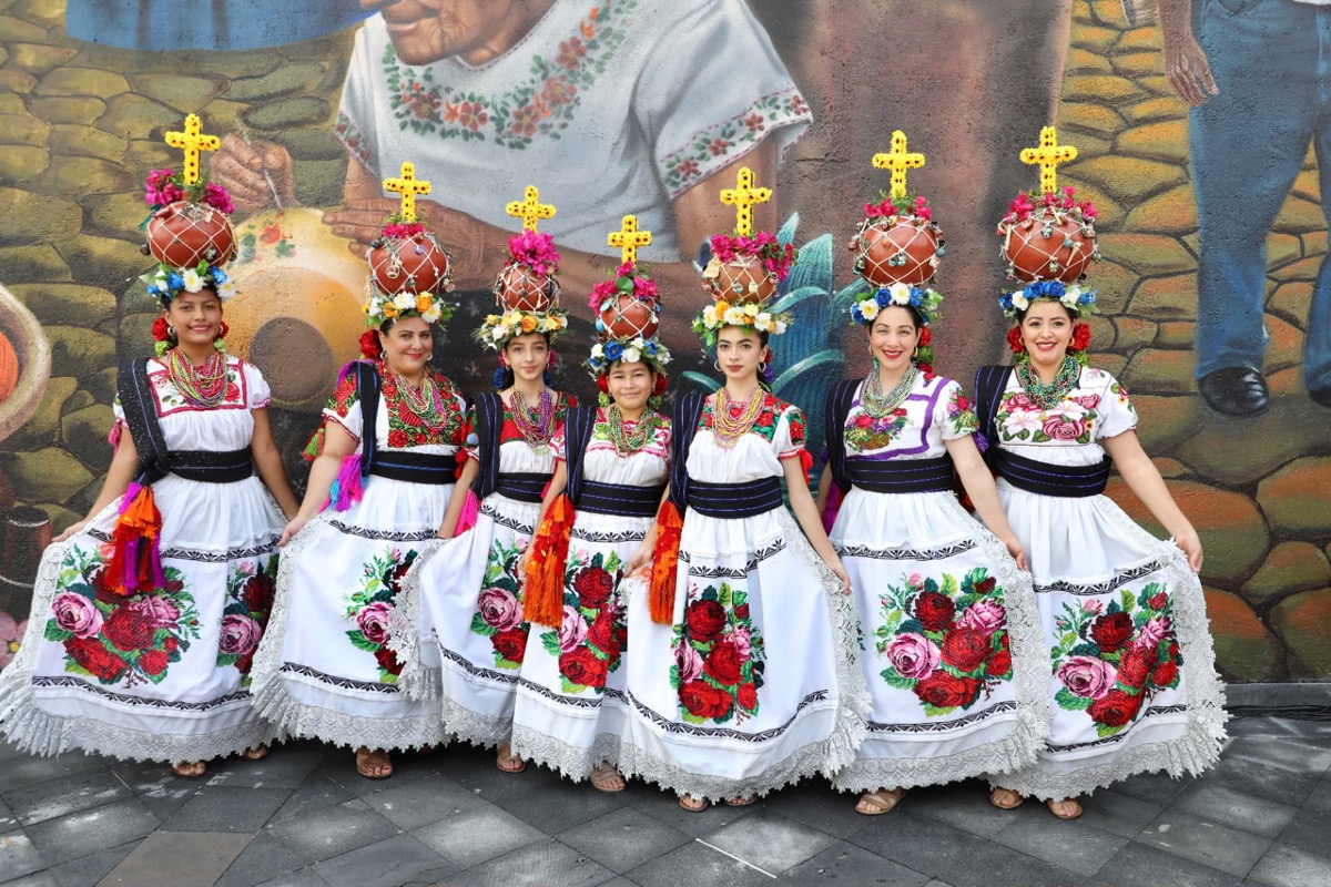 Siete mujeres con trajes tradicionales blancos con flores y jarrones decorados en la cabeza posan frente a un mural colorido.