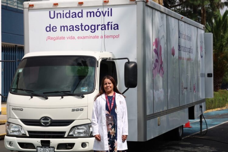 Una mujer con bata blanca está de pie frente a una unidad móvil de mastografía estacionada.