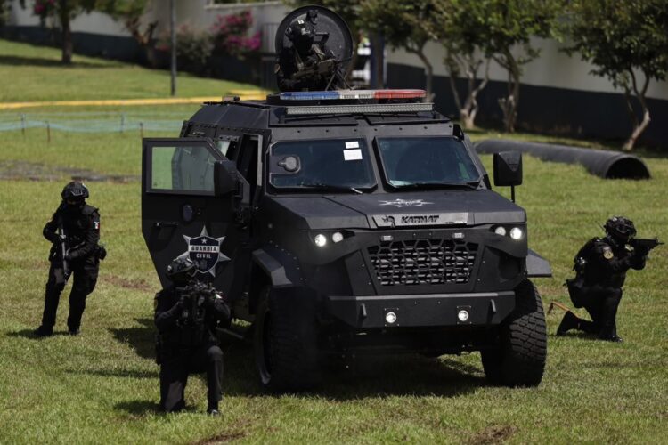 Un vehículo blindado negro con soldados armados alrededor realiza un ejercicio en un campo de césped.
