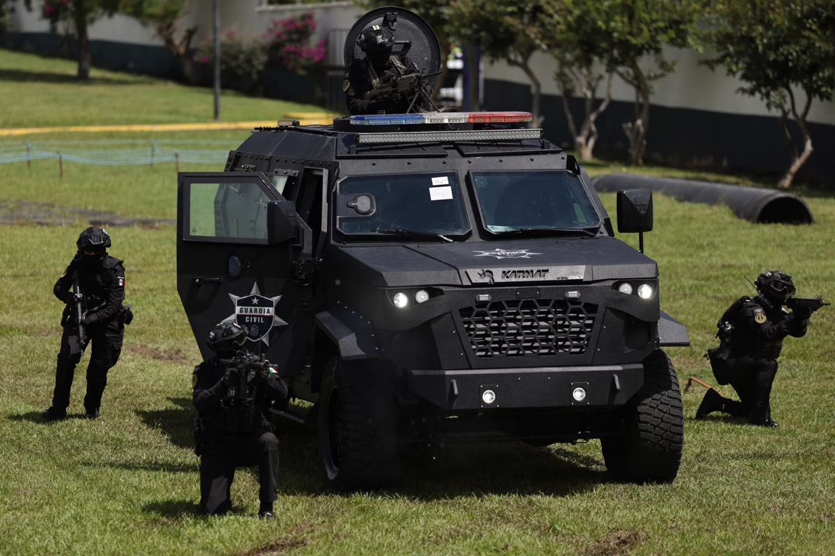 Un vehículo blindado negro con soldados armados alrededor realiza un ejercicio en un campo de césped.