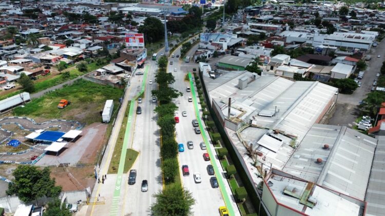 Vista aérea de una calle urbana con tráfico moderado, ciclovías verdes y edificios residenciales e industriales a los lados.