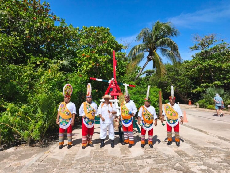 Seis hombres con trajes tradicionales coloridos posan frente a un mástil rojo en un entorno tropical con palmeras.