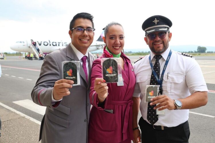 Tres empleados de aerolínea sonríen en la pista mostrando recuerdos de Michoacán, con un avión de Volaris al fondo.